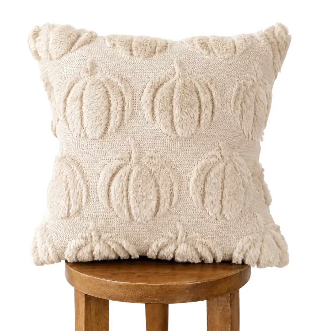 A square cream throw pillow with a raised, textured pumpkin pattern, resting on a wooden stool against a solid white background.