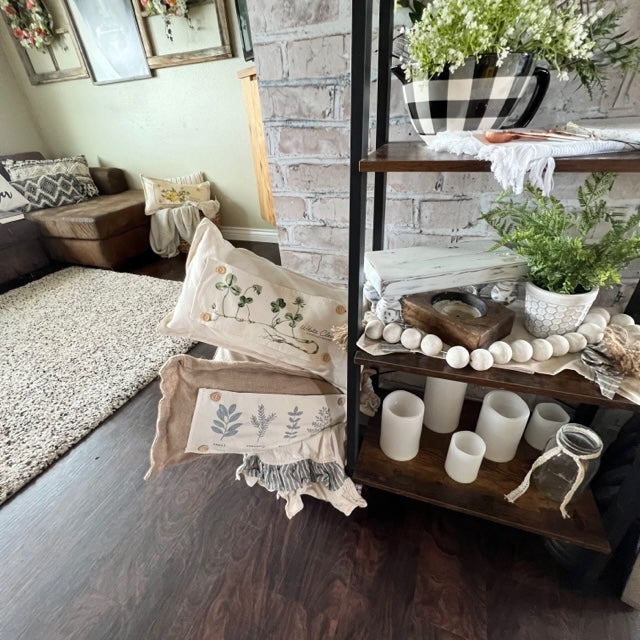 Two decorative throw pillows in a natural beige linen fabric are styled on a dark wood shelf in a cozy living room setting. The top pillow features a green botanical leaf pattern, and the bottom pillow has a gray botanical leaf pattern with a fringed edge.