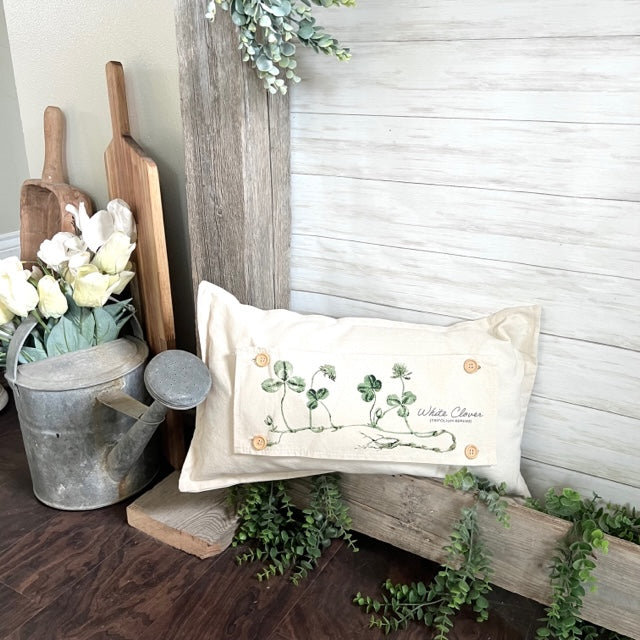 A rectangular cream throw pillow with a St. Patrick's Day embroidered design featuring shamrocks and the text 'White Clover', styled on a rustic wooden bench in a farmhouse-themed room setting.