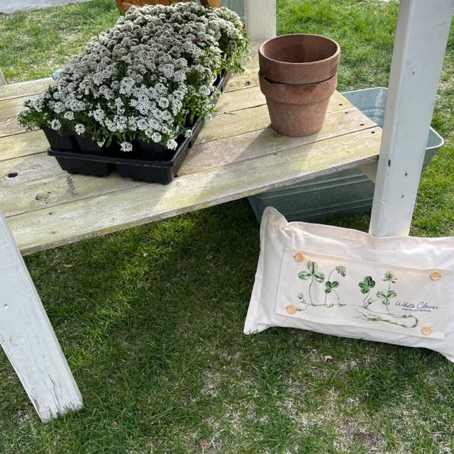 A cream-colored decorative throw pillow with a botanical illustration of white flowers and the text 'White Clover' resting on green grass next to a wooden garden table.