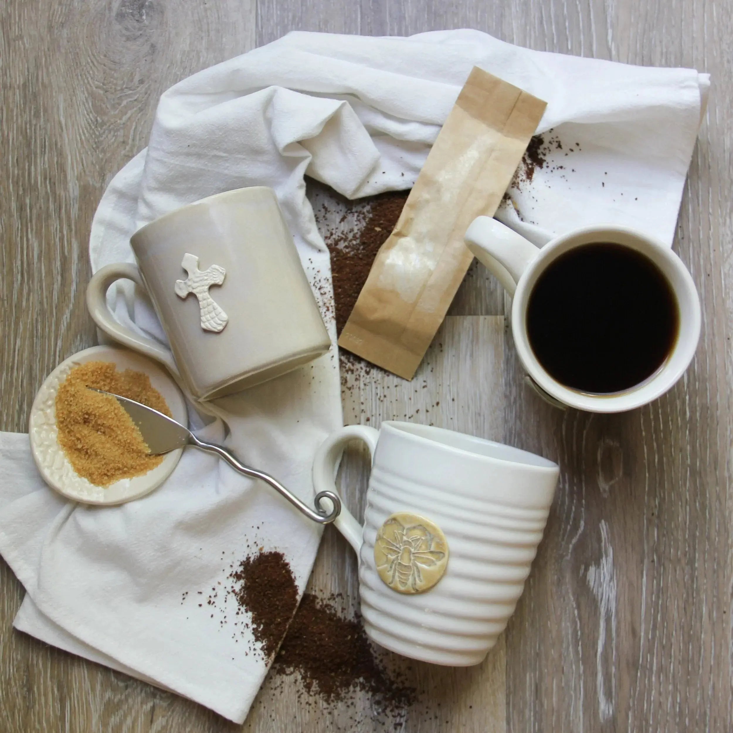 Cozy farmhouse coffee mugs with cross and bee designs, brown sugar, and coffee grounds on rustic wood
