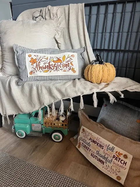A styled autumn home decor scene featuring a striped throw pillow with 'Thanksgiving' embroidered in orange and yellow, resting on a cream fringed blanket next to a small orange pumpkin. A teal toy truck sits below on a wood floor, and a burlap pillow with fall-themed text lies on the floor nearby.