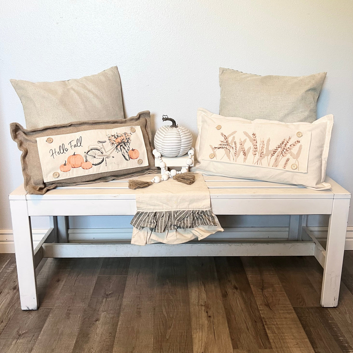 Two decorative throw pillows styled on a white wooden bench against a white wall, with a small striped pumpkin and a folded fringed throw blanket. The left pillow is brown with a 'Hello Fall' autumn design, and the right pillow is cream with a wheat design.