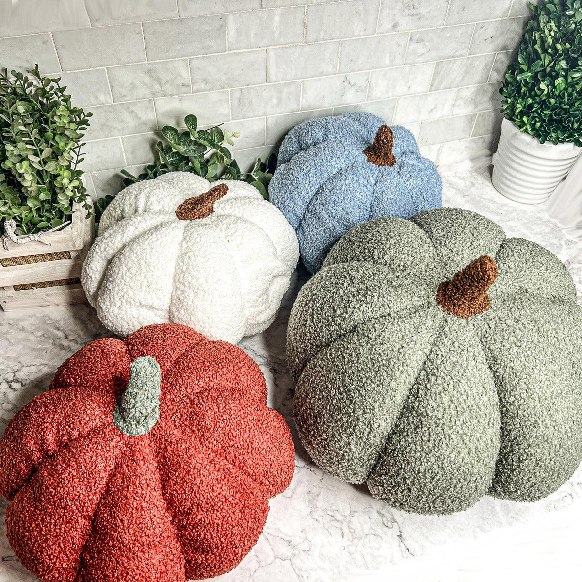 Four textured pumpkins in white, blue, red, and green on a light surface with plants in the background.