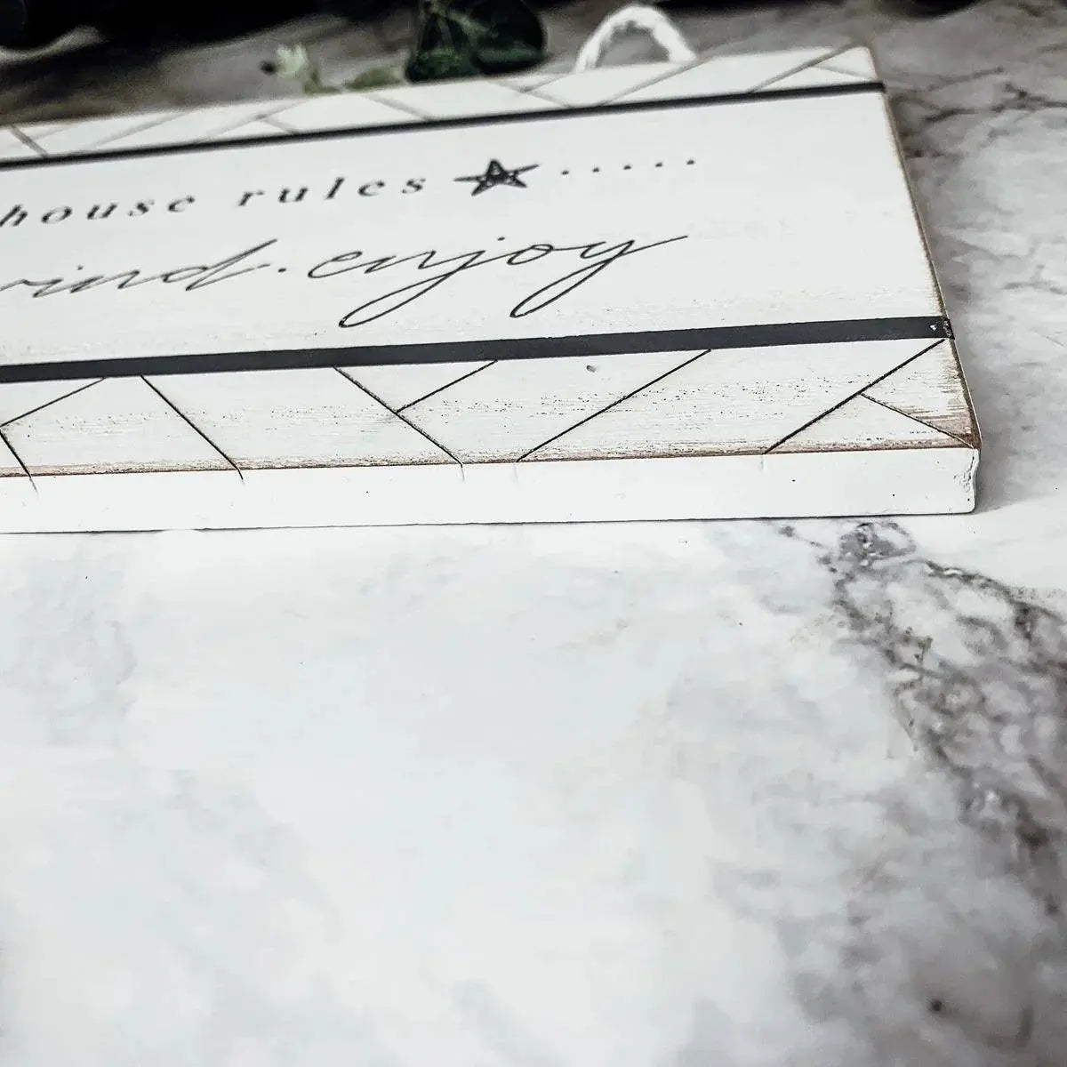 White farmhouse-style wooden sign with 'house rules' text and geometric patterns on marble surface