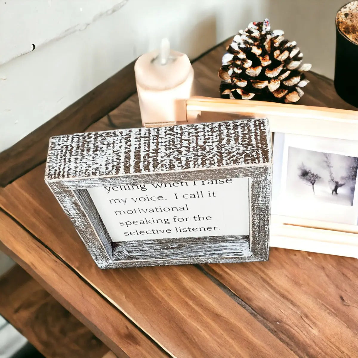 Decorative box with text on a wooden surface with a candle and pine cone in the background.