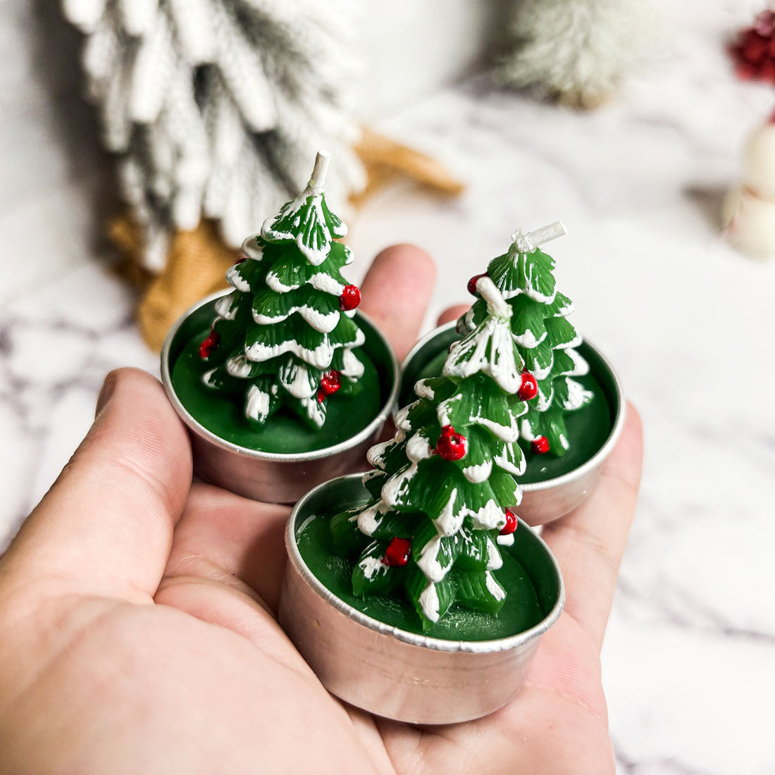 Three Christmas tree-shaped candles held in a hand with a blurred festive background.