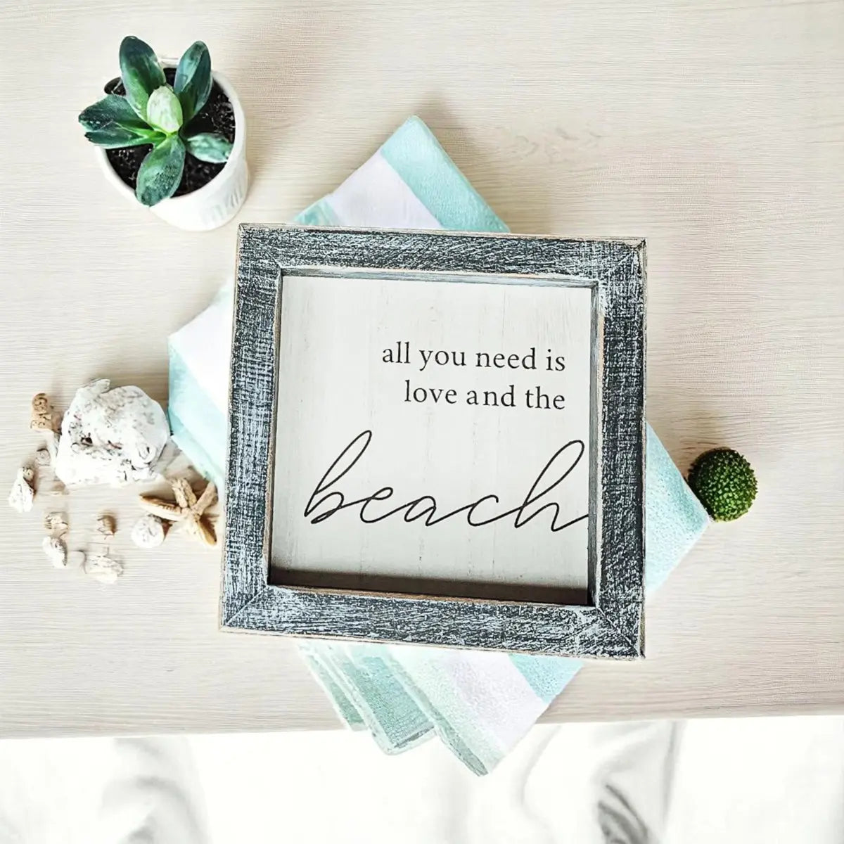 Framed sign with 'all you need is love and the beach' text on a decorative surface with plants and shells.