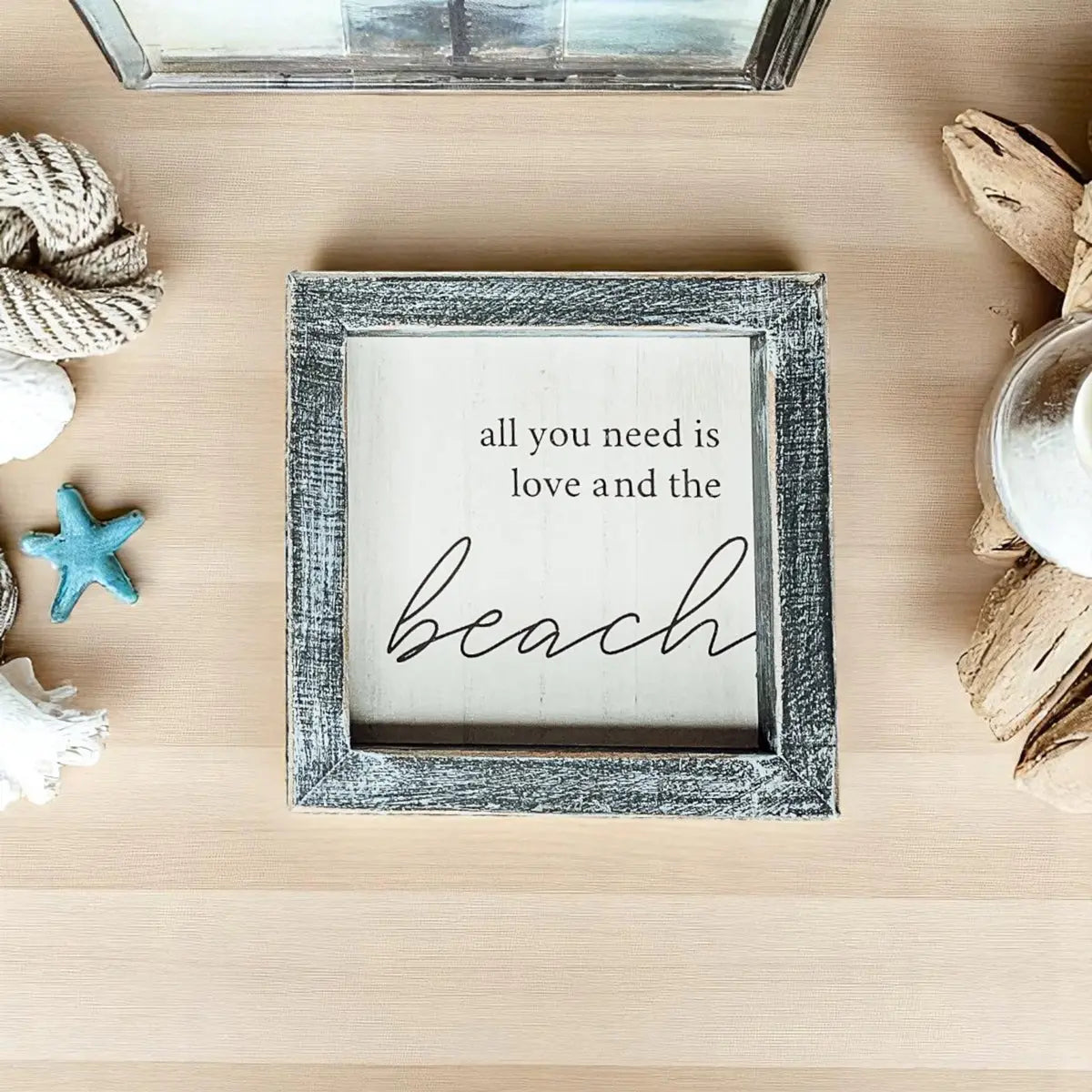 Framed sign with 'all you need is love and the beach' text on a wooden surface with beach-themed decor.