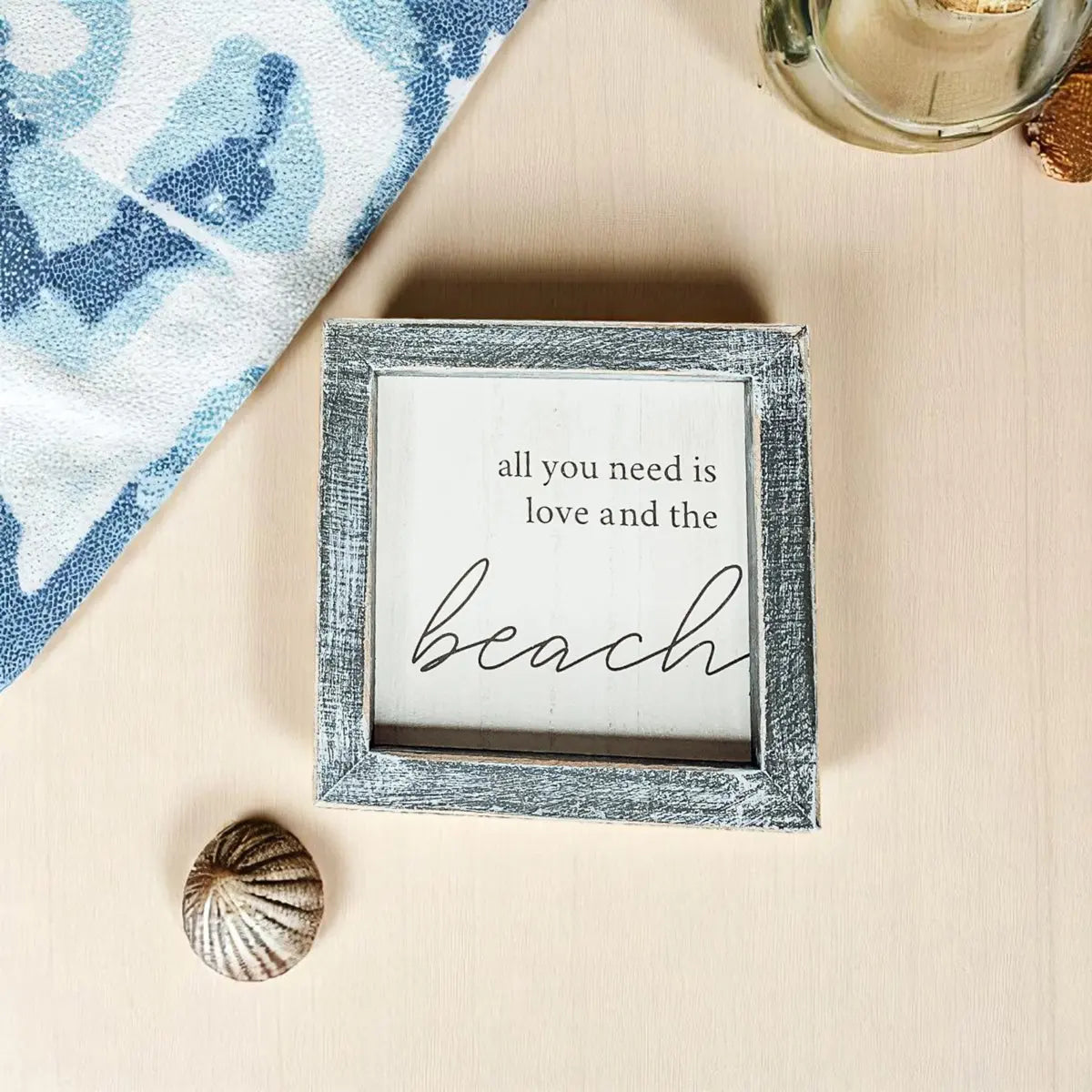 Decorative sign with 'all you need is love and the beach' text on a sandy surface with a shell and towel.
