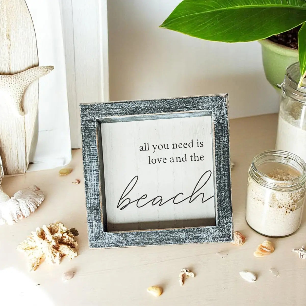 Decorative sign with 'all you need is love and the beach' text in a rustic frame on a beach-themed setting.