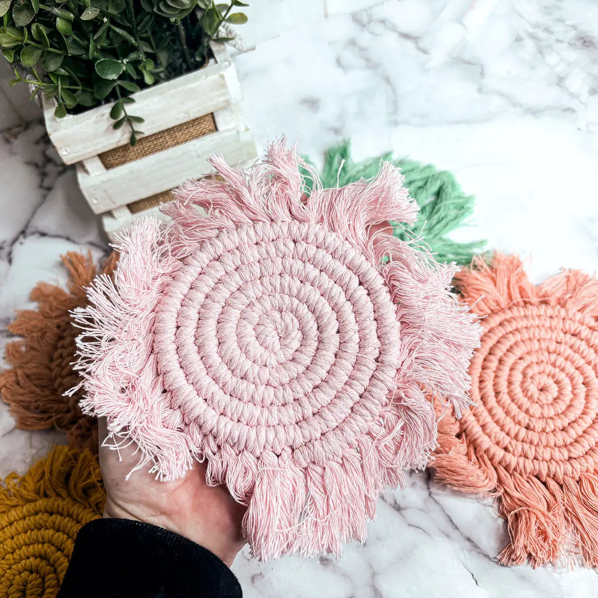 Pink macrame coaster held by a hand on a marble surface with other coasters in the background.