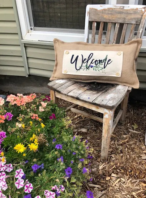 A rustic burlap welcome pillow with a cream label reading 'Welcome' in black script and small green leaves, resting on a weathered wooden chair outdoors next to a flower bed.