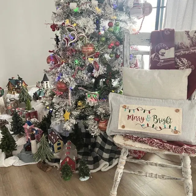 A decorated Christmas tree in a living room setting next to a distressed white rocking chair with a 'Merry & Bright' throw pillow, surrounded by miniature houses and gifts on the floor.