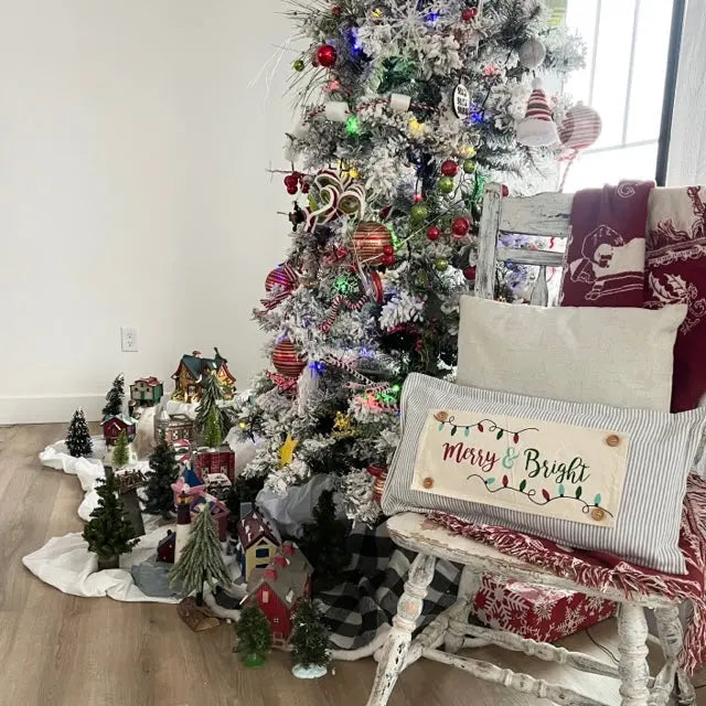 A cozy holiday living room scene featuring a decorated white flocked Christmas tree, a distressed white rocking chair with red and white patterned cushions, and a square throw pillow that reads 'Merry & Bright' with colorful lights. Small Christmas village figurines are arranged on the floor beside the tree.