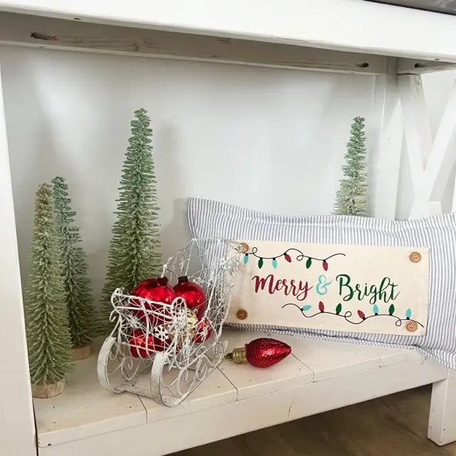 A rectangular holiday throw pillow with a cream center panel and blue-and-white striped sides, printed with the words 'Merry & Bright' and colorful Christmas lights, displayed on a white shelf beside miniature trees, a white wire sleigh with red ornaments, and a loose red ornament.