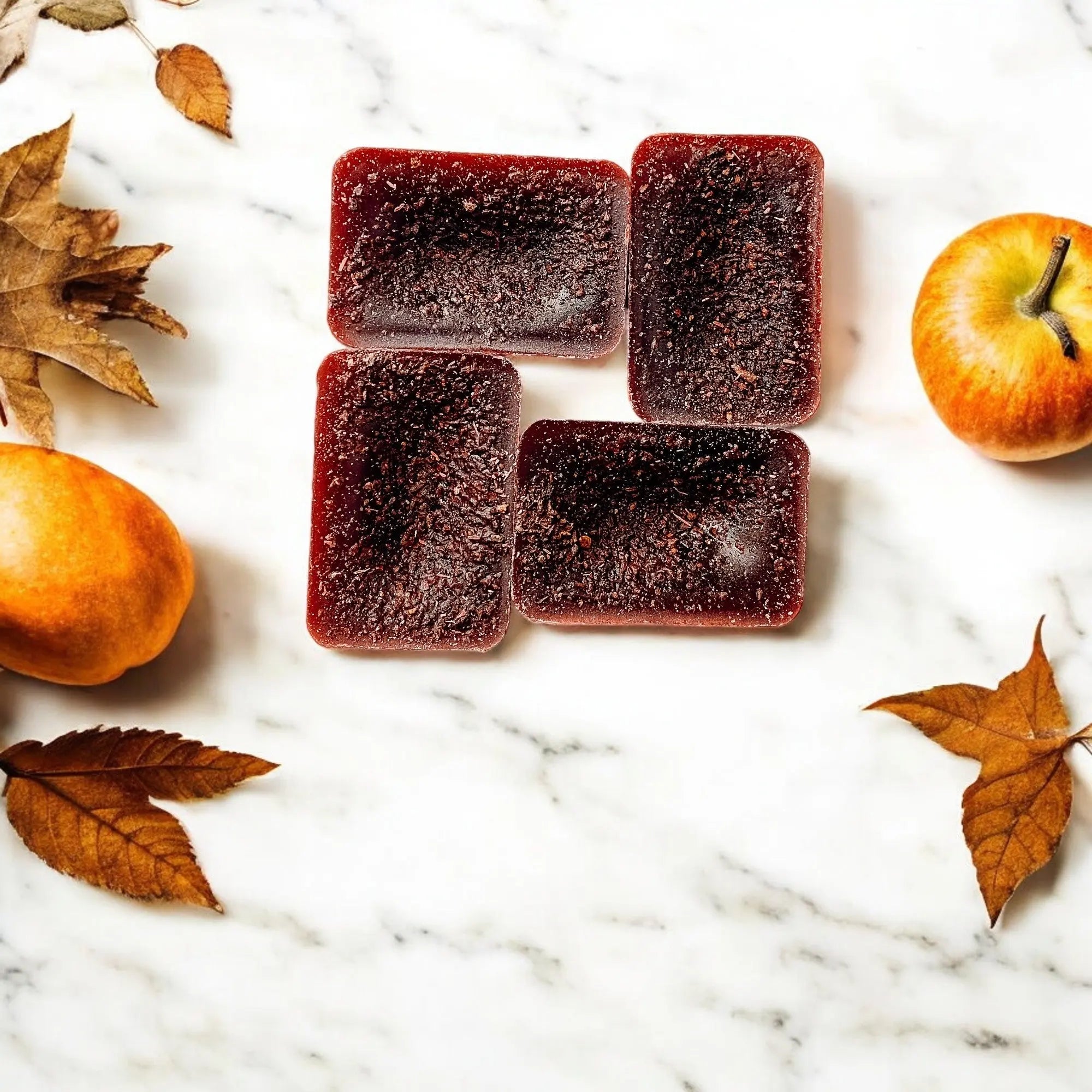Five square pieces of dark red jelly on a marble surface with apples and leaves.