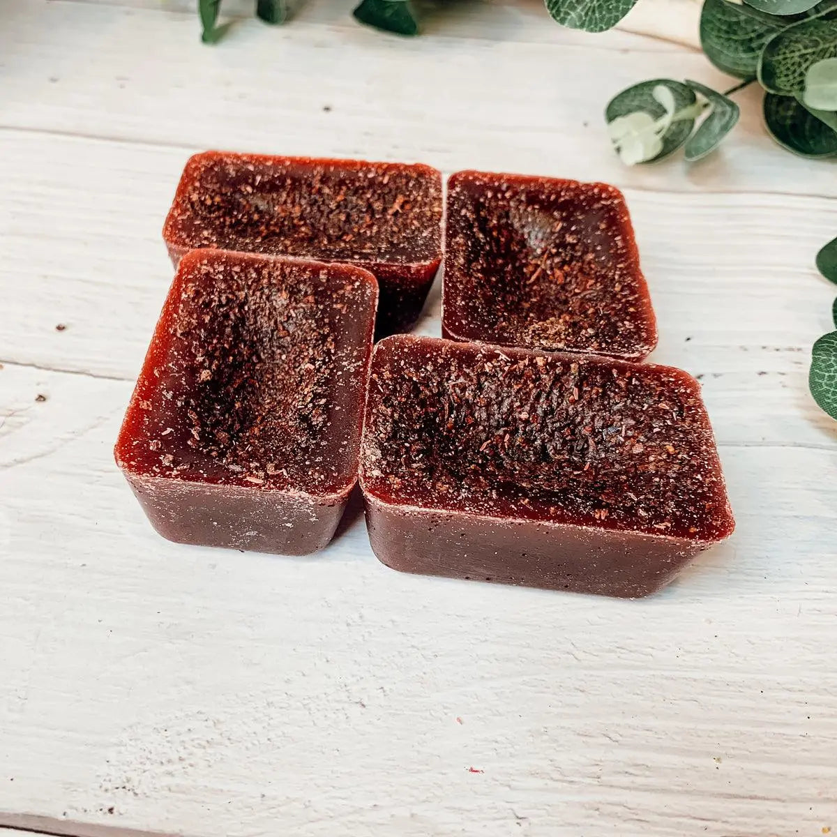 Four square pieces of dark red soap on a light wooden surface with green leaves in the background.