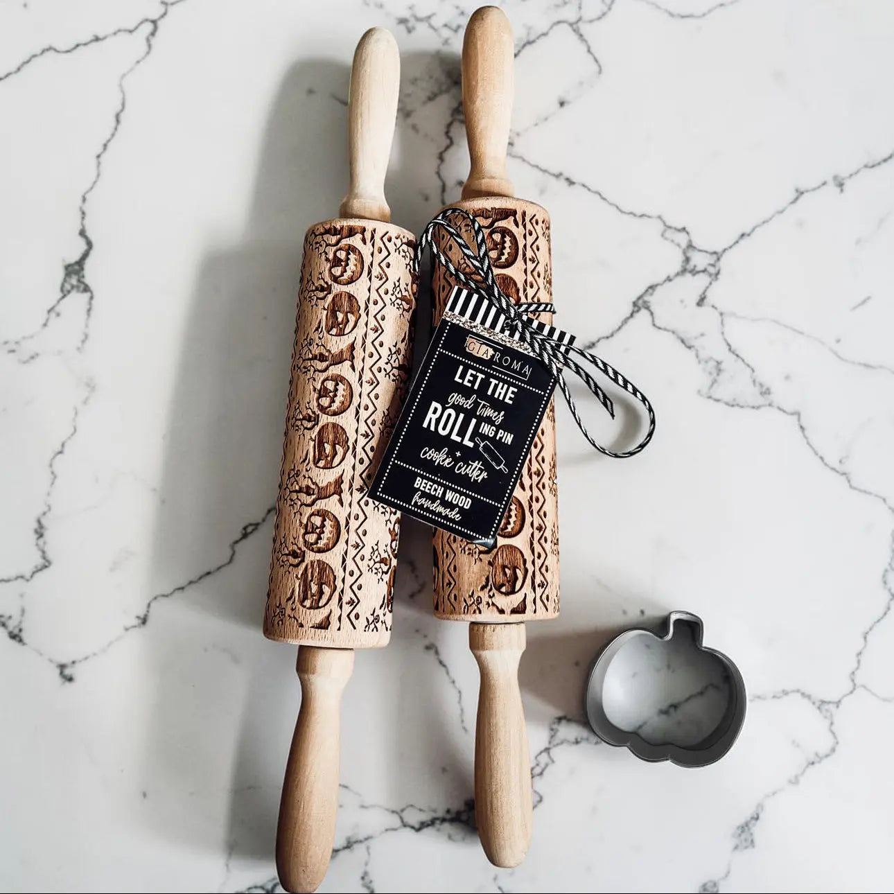 Two wooden rolling pins with embossed designs on a marble surface, accompanied by a tag and a small round cookie cutter.