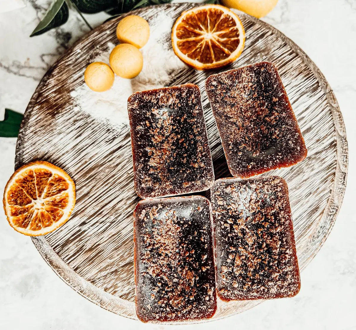 Four dark brown square pastries on a round wooden board with sliced oranges and lemons.