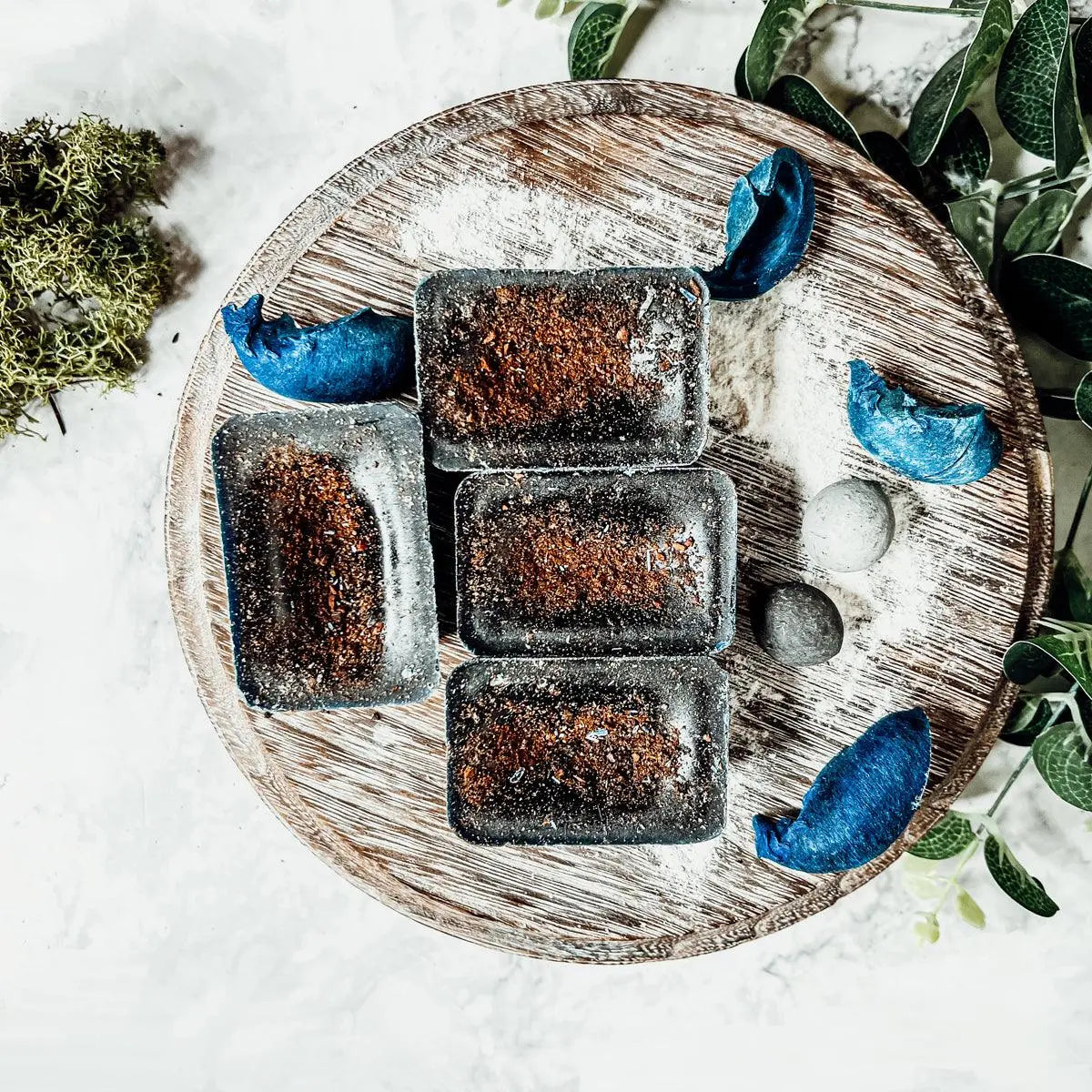 Five dark brown rectangular pastries on a wooden plate with decorative blue leaves and greenery.