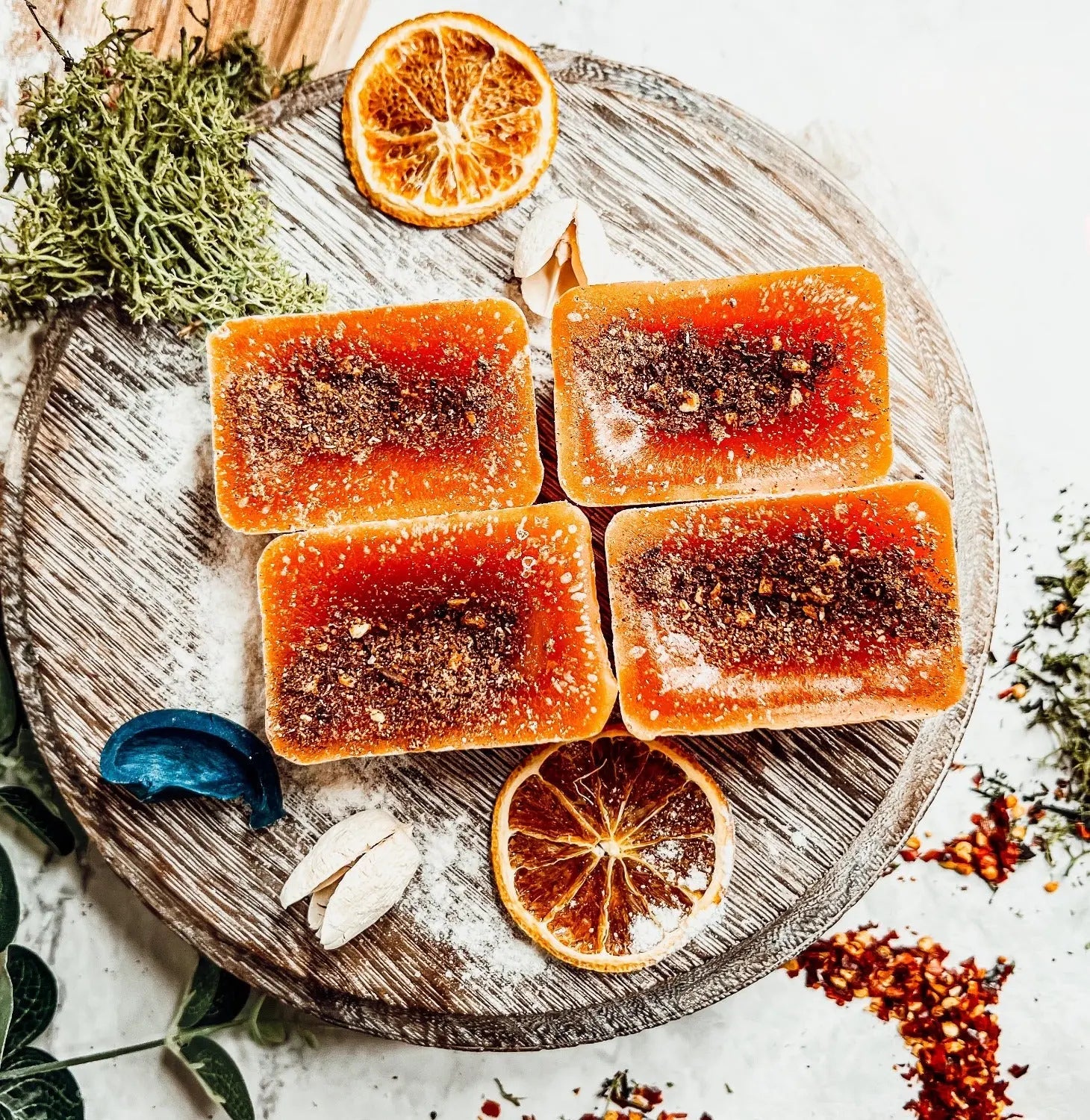 Four orange-colored soap bars with specks on a wooden board, surrounded by dried oranges and herbs.