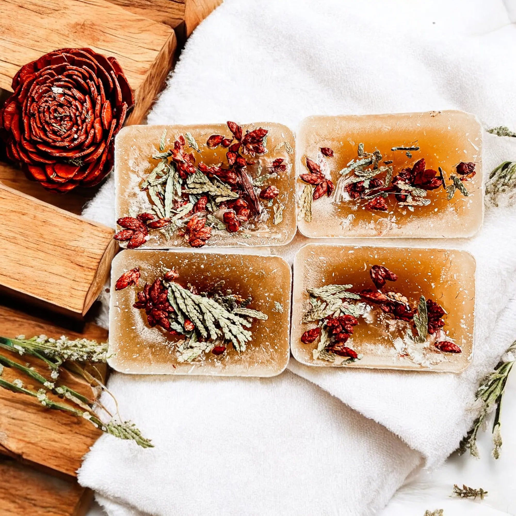Five rectangular soap bars with natural ingredients on a white towel with wooden blocks and dried flowers.