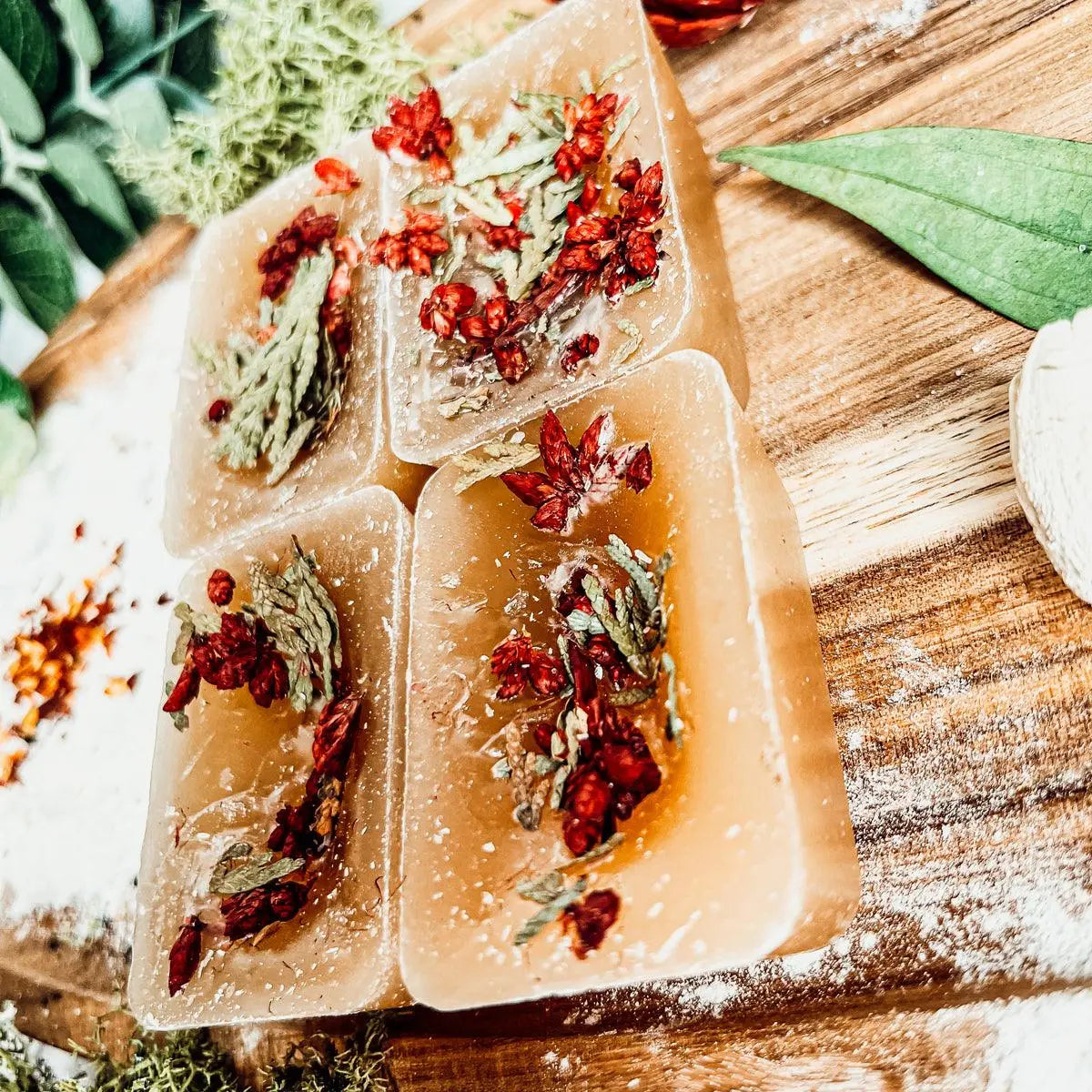 Transparent soap bars with red and green herbs on a wooden surface with leaves.