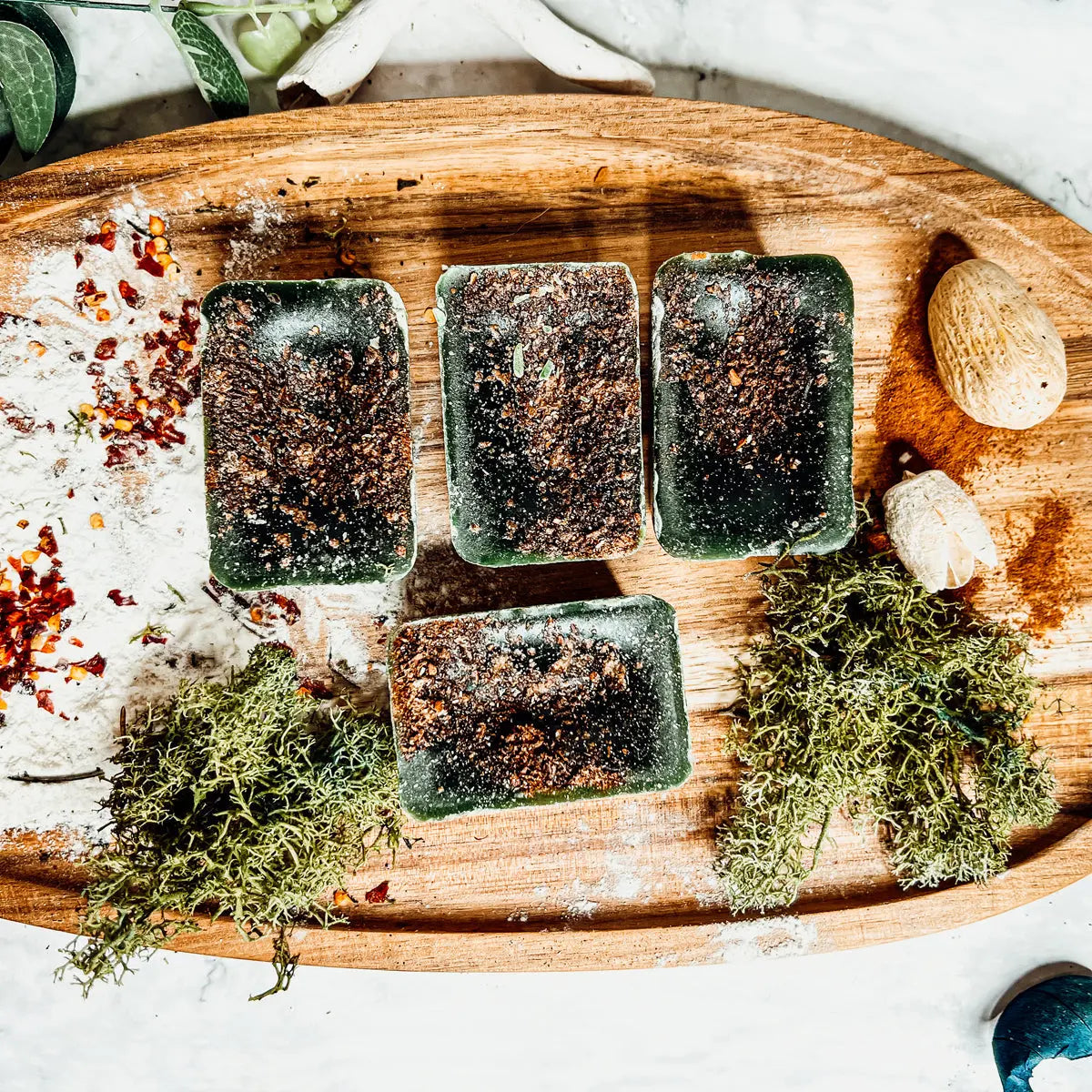 Five dark green soap bars on a wooden cutting board with herbs and spices.