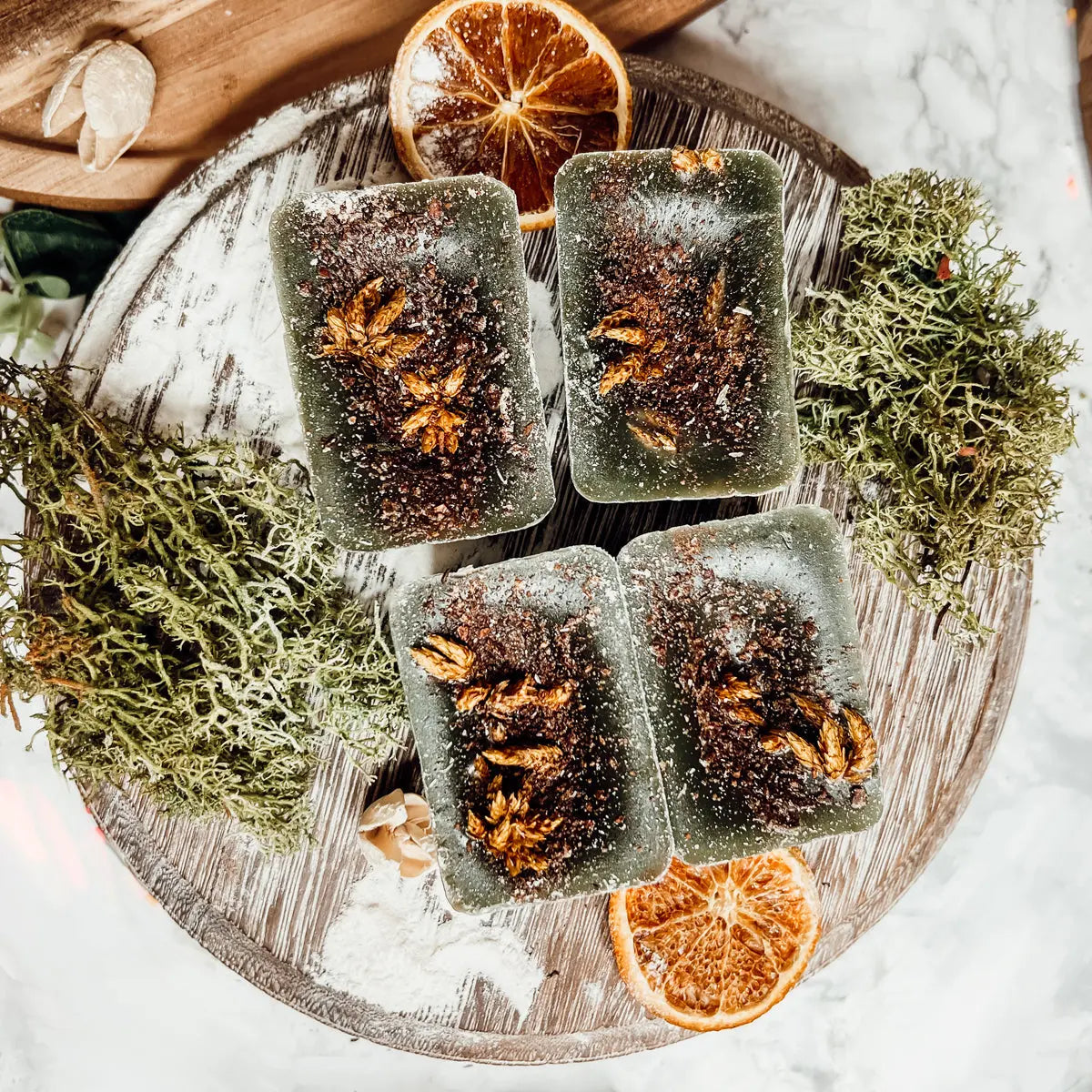 Five rectangular soap bars with botanical elements on a wooden board with dried oranges and moss.
