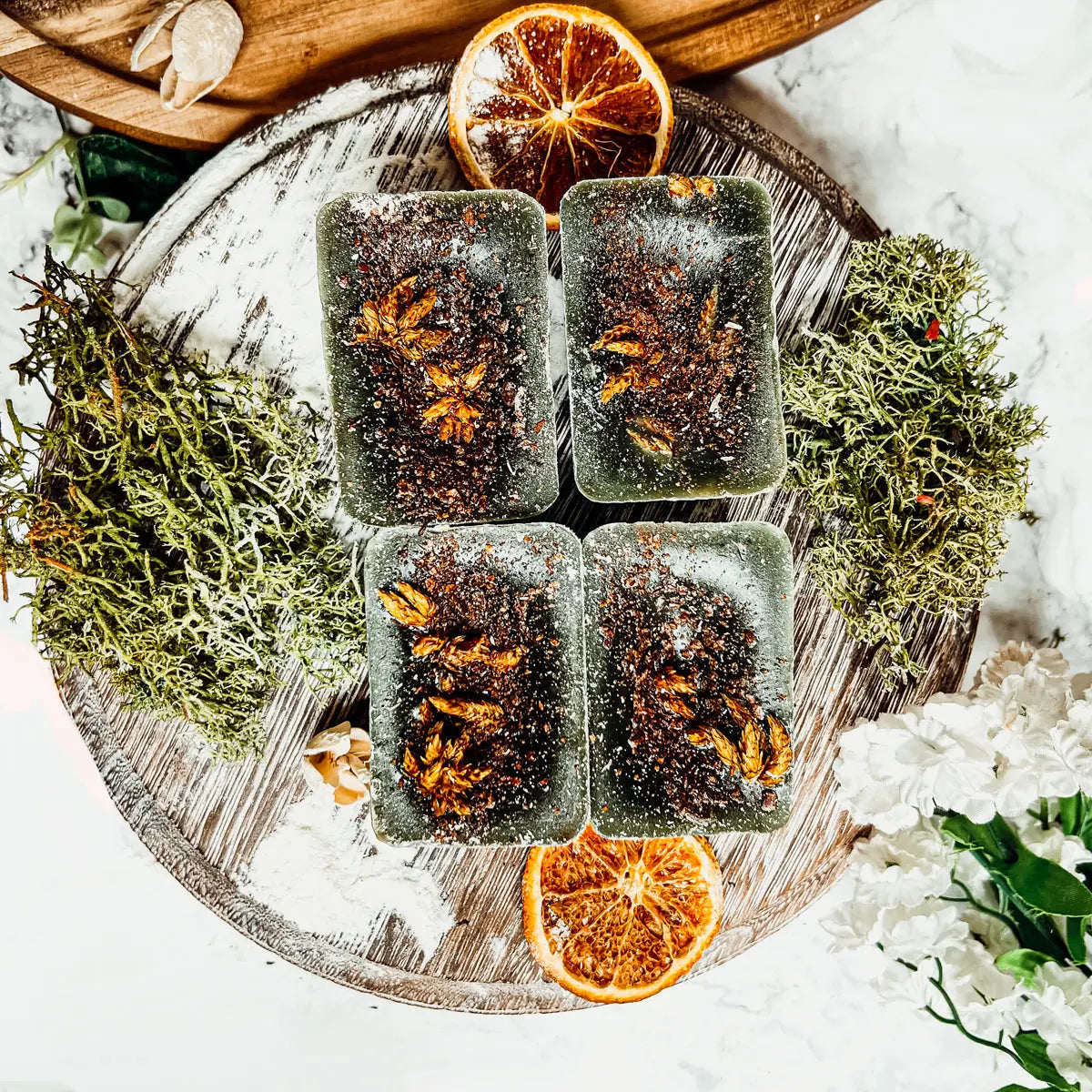 Four rectangular soap bars with leaf patterns on a wooden board with dried oranges and greenery.