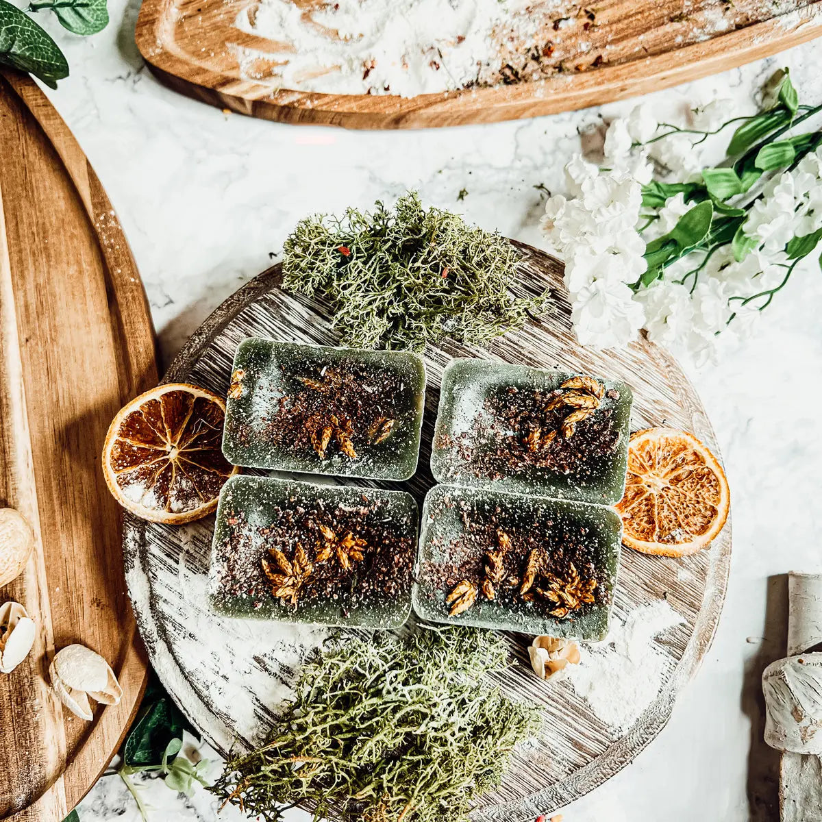 Dark green soap bars with visible herbs on a wooden board with dried oranges and greenery.