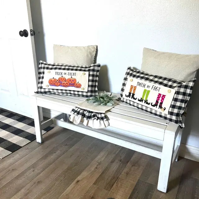 A white wooden bench styled with two black-and-white buffalo plaid throw pillows featuring Halloween-themed designs, including pumpkins and witch boots, set on a wood floor against a white wall.