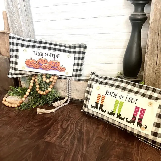 Two rectangular black-and-white buffalo plaid decorative pillows displayed on a dark wood floor in a rustic fall-themed setting. One pillow reads "TRICK OR TREAT" with carved pumpkins, and the other reads "SMELL MY FEET" with witch boots.