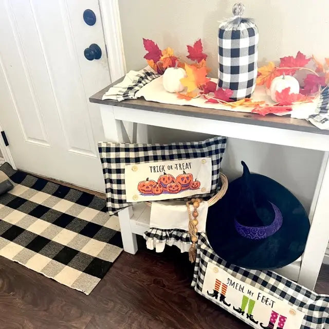A styled fall entryway scene with a white console table holding a buffalo plaid pillow, white pumpkins, red and orange autumn leaves, a black witch hat, and another plaid decorative pillow on a shelf.
