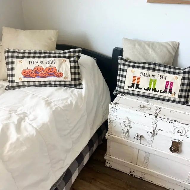 Two decorative rectangular throw pillows in a black-and-white buffalo plaid pattern are styled on a bed and a distressed white trunk in a bedroom. The pillow on the bed reads 'TRICK OR TREAT' with orange jack-o'-lantern illustrations, and the pillow on the trunk reads 'SMELL MY FEET' above black cat silhouettes and colorful striped socks.