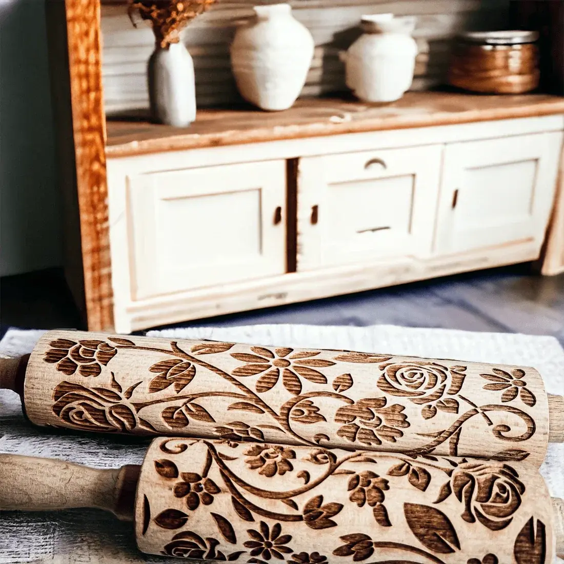 Two wooden embossing rolling pins with floral patterns on a surface, with a wooden cabinet and decorative items in the background.