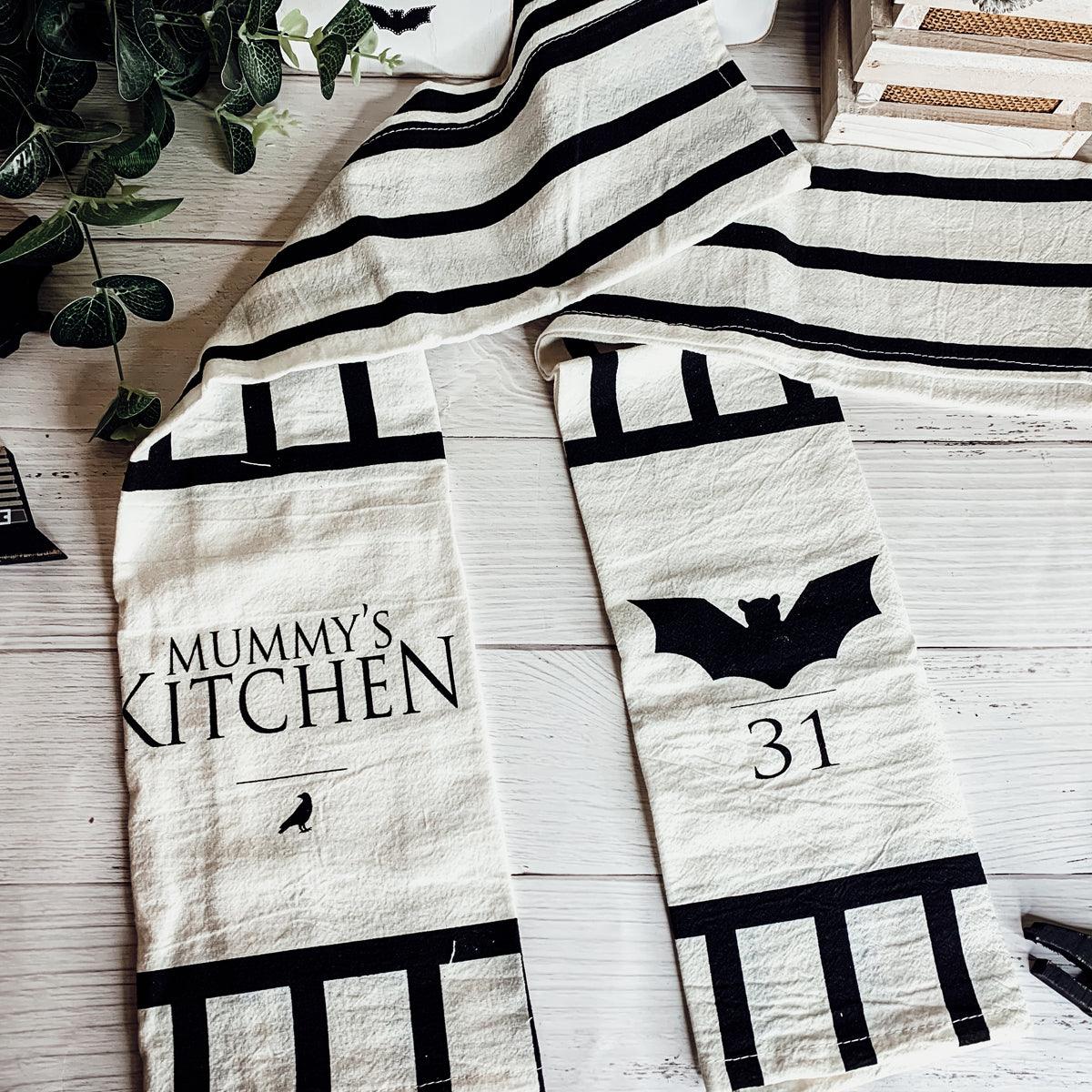 Halloween-themed kitchen towels with text and bat designs on a wooden surface.