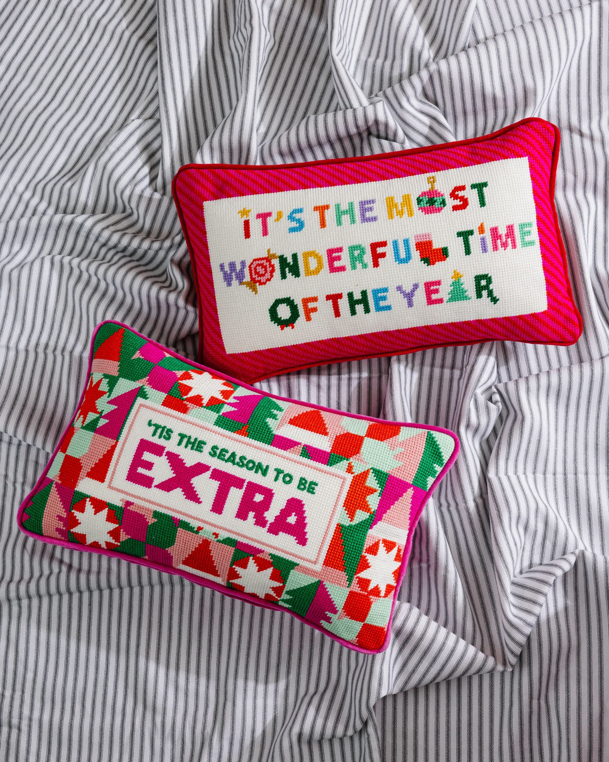 Two rectangular Christmas throw pillows on a gray and white striped bedsheet background. The top pillow has a red border and a white center with colorful embroidered text reading 'IT'S THE MOST WONDERFUL TIME OF THE YEAR'.