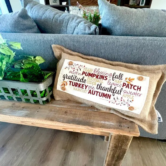 A rectangular burlap throw pillow with an autumn-themed word cloud design reading 'Blessing from above, PUMPKINS, fall leaves, PATCH sweater, THANK YOU turkey, thankful autumn pie' is styled on a rustic wooden bench in front of a gray sofa.