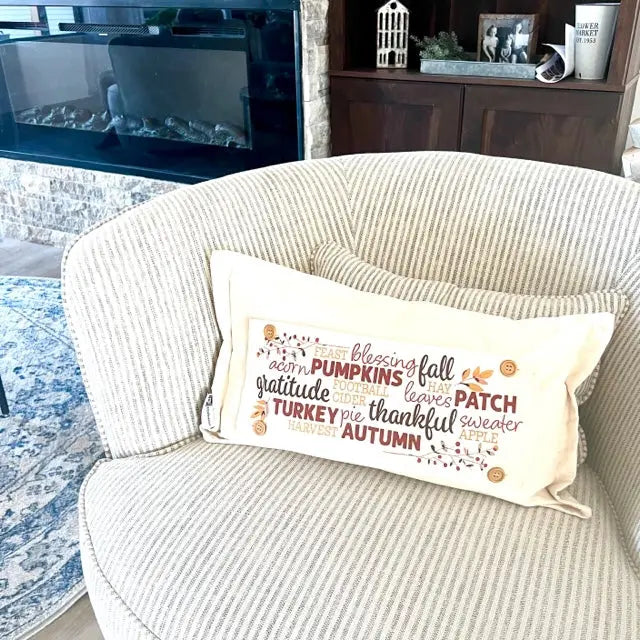 A cream-colored lumbar throw pillow with a fall-themed text design, resting on a striped beige armchair in a cozy living room setting.