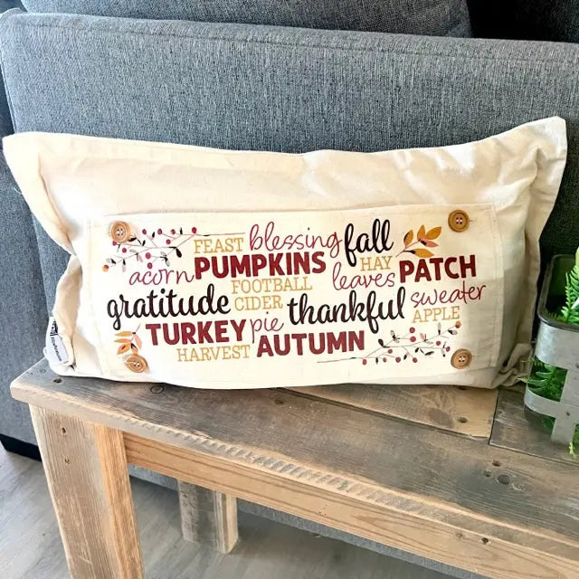 An off-white rectangular throw pillow with a warm autumn-themed word collage print, resting on a rustic wooden table in front of a gray fabric sofa.