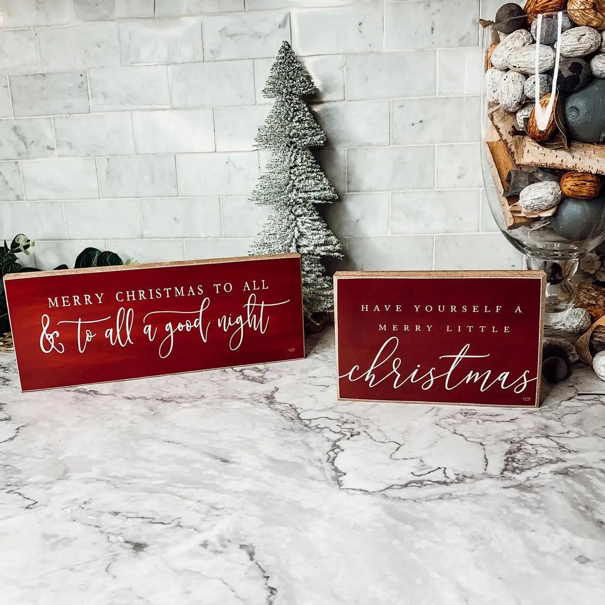 Two red Christmas signs with white text on a marble surface with decorative items.