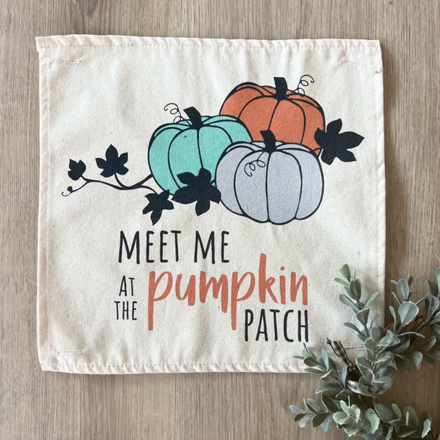 A square cream fabric napkin or small towel laid flat on a light wood surface, printed with three illustrated pumpkins in teal, orange, and pale gray, black leaves, and the phrase “MEET ME AT THE pumpkin PATCH.” A small greenery sprig appears at the lower right corner.