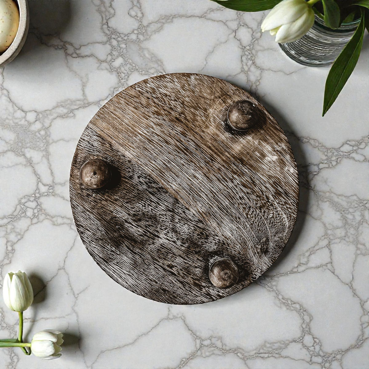 Round wooden cutting board with three knobs on a marble surface with tulips.