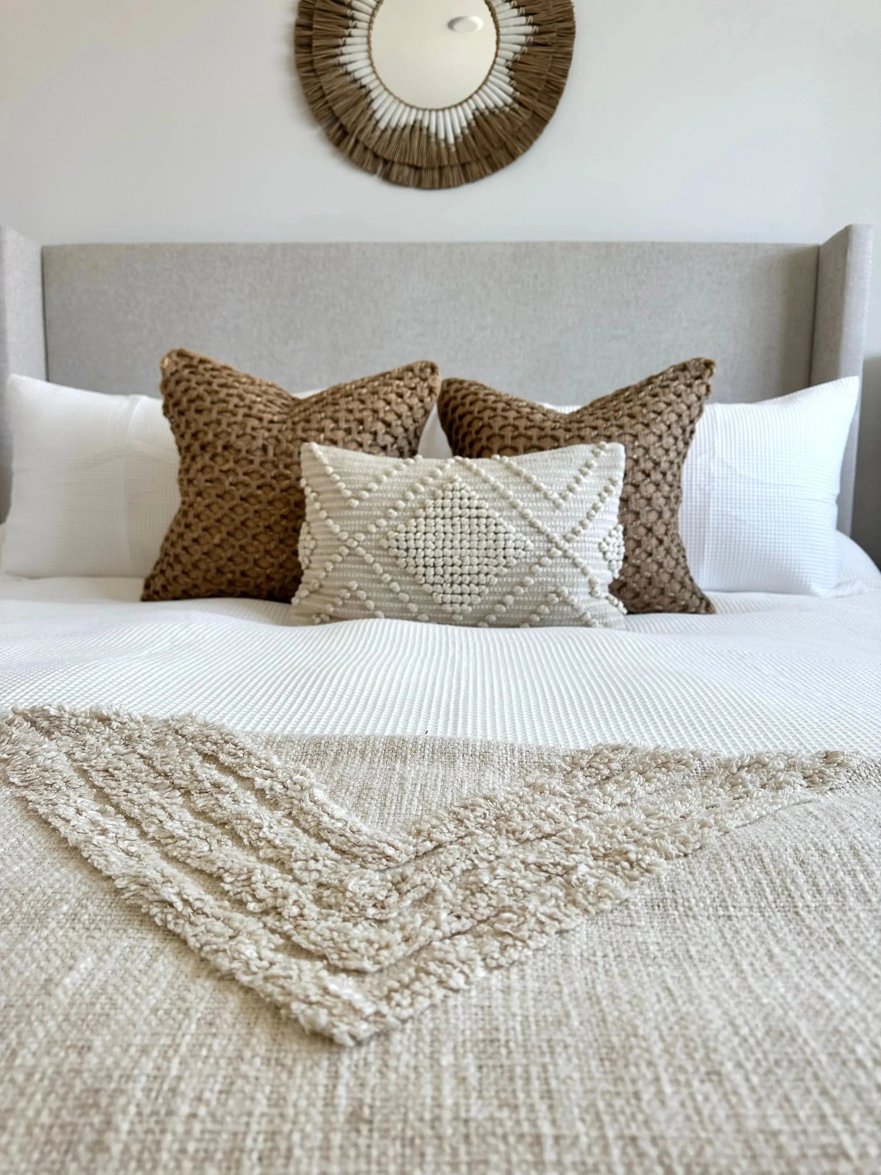 Cozy farmhouse bedroom with textured throw pillows, woven blanket, and sunburst mirror above bed