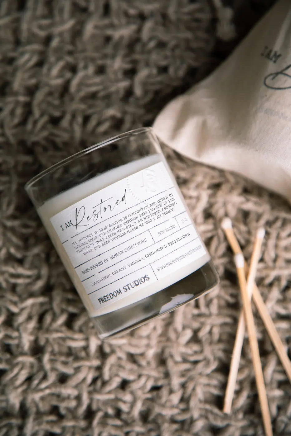 I Am Restored soy candle with cozy farmhouse aesthetic, hand-poured by women survivors, on textured knit blanket