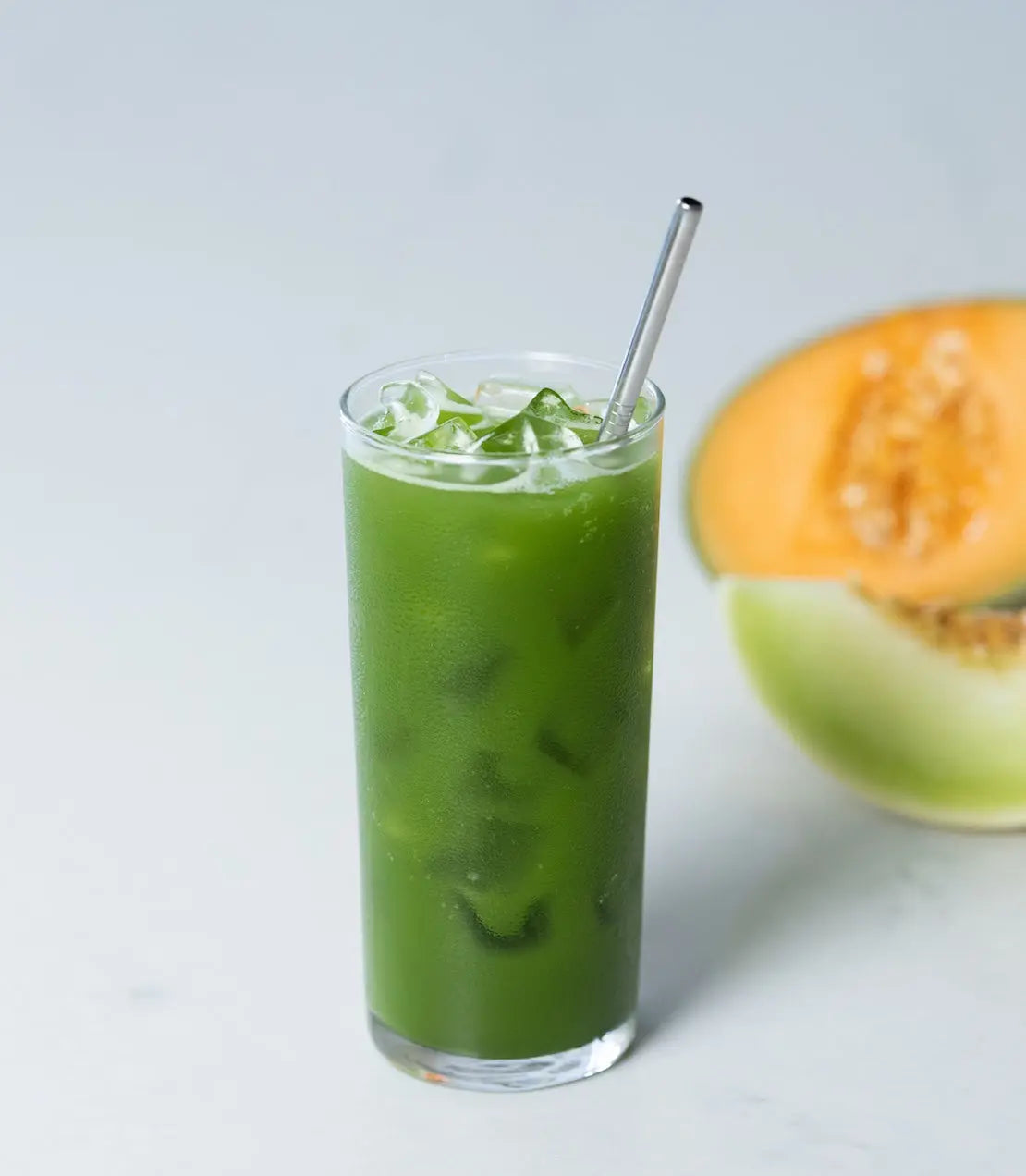 Iced green melon juice in tall glass with metal straw, fresh cantaloupe slice beside