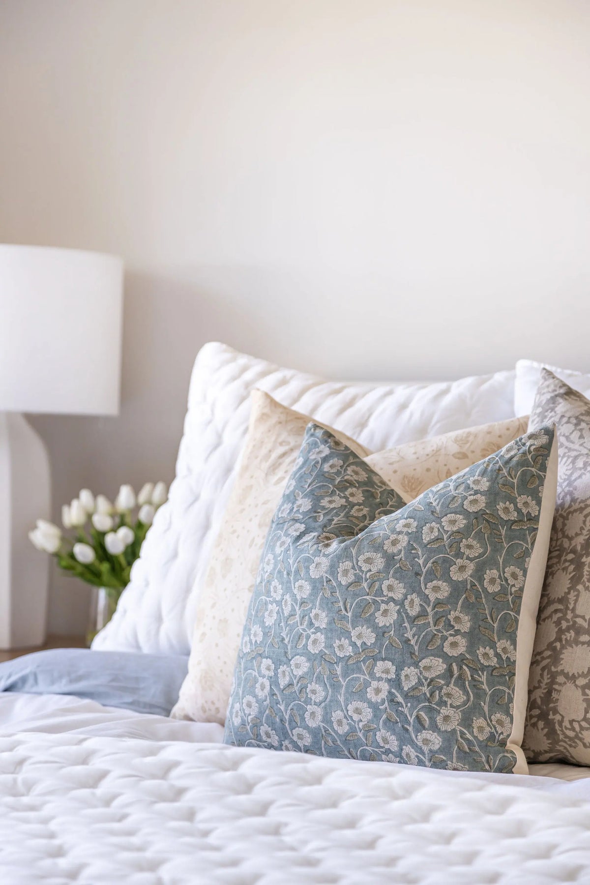 Decorative pillows on a bed with a lamp and flowers in the background
