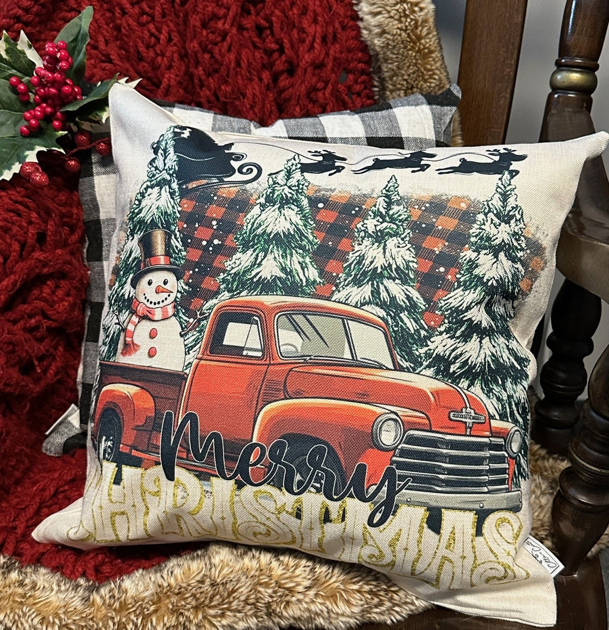 A square Christmas throw pillow featuring a vintage red pickup truck carrying a snowman, set against snowy pine trees and a plaid holiday background with 'Merry Christmas' text. The pillow is styled on a red knit blanket and plaid pillow on a wooden chair.
